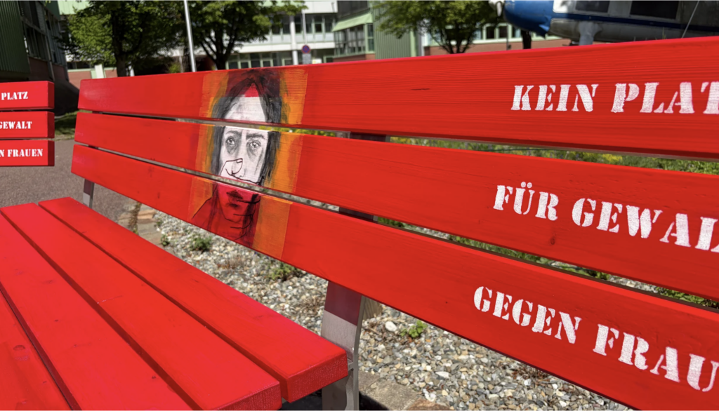 Eine knallrote Parkbank mit einem gemalten Frauengesicht auf der Lehne.